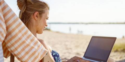 Frau sitzt am Strand und arbeitet an ihren Laptop