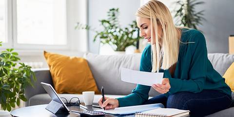 Lächelnde Frau, die Rechnungen analysiert und Steuerunterlagen ausfüllt, während sie zu Hause auf dem Sofa sitzt