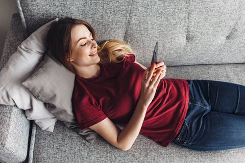 Frau mit rotem Shirt liegt auf der Couch und schaut in ihr Handy