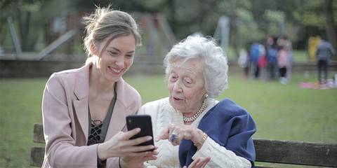 Junge Frau sitzt mit einer älteren Frau auf einer Parkbank und zeigt ihr etwas auf dem Smartphone - Artikelbild von Lexware