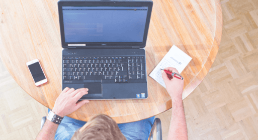 Vogelperspektive: Person sitzt am Tisch vor einem Laptop und schreibt etwas in sein Notizblock